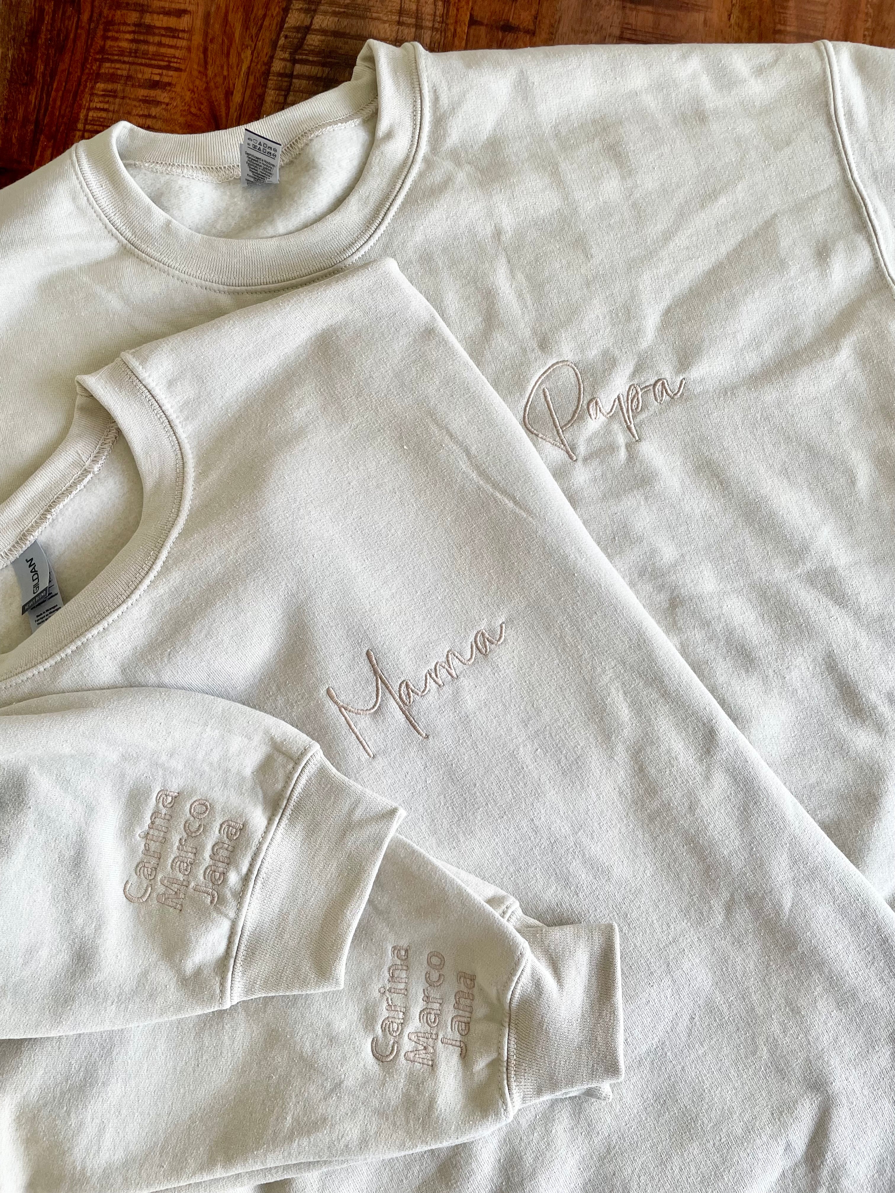 Zwei personalisierte und bestickte Sweater ohne Kapuze, worauf Mama und Papa stehen. Am Ärmel sind die Namen der gemeinsamen Kinder gestickt.
