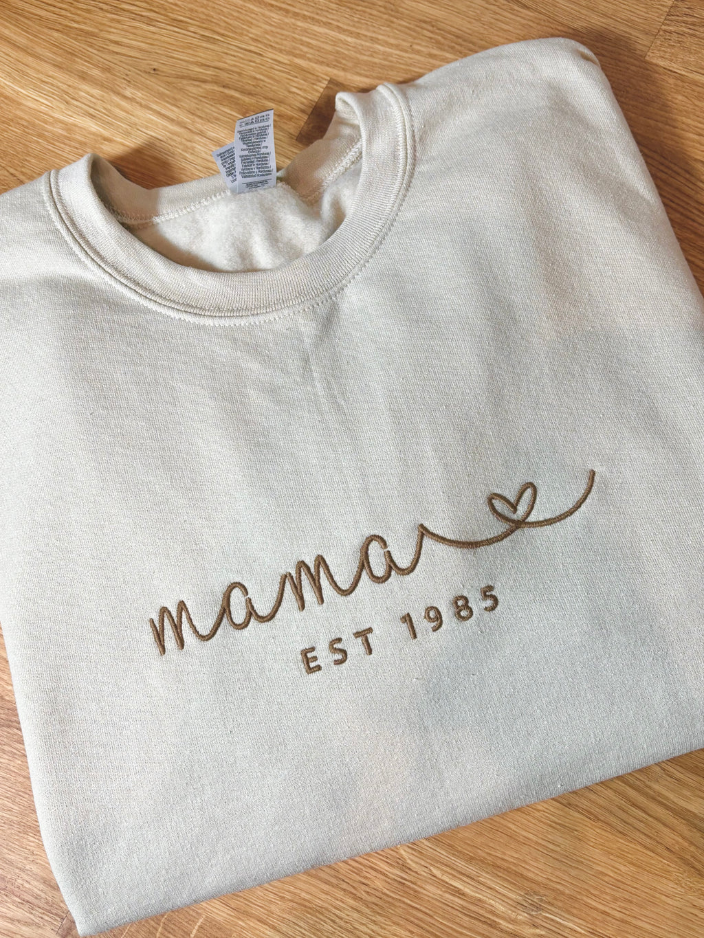 Ein personalisierter beiger Sweater mit einem braunen Stick "mama est. 1985§" mit einem Herz.