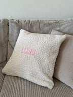 Ein besticktes Kissen aus Boucle Stoff mit einem rosa Wunschtext "Luisa".