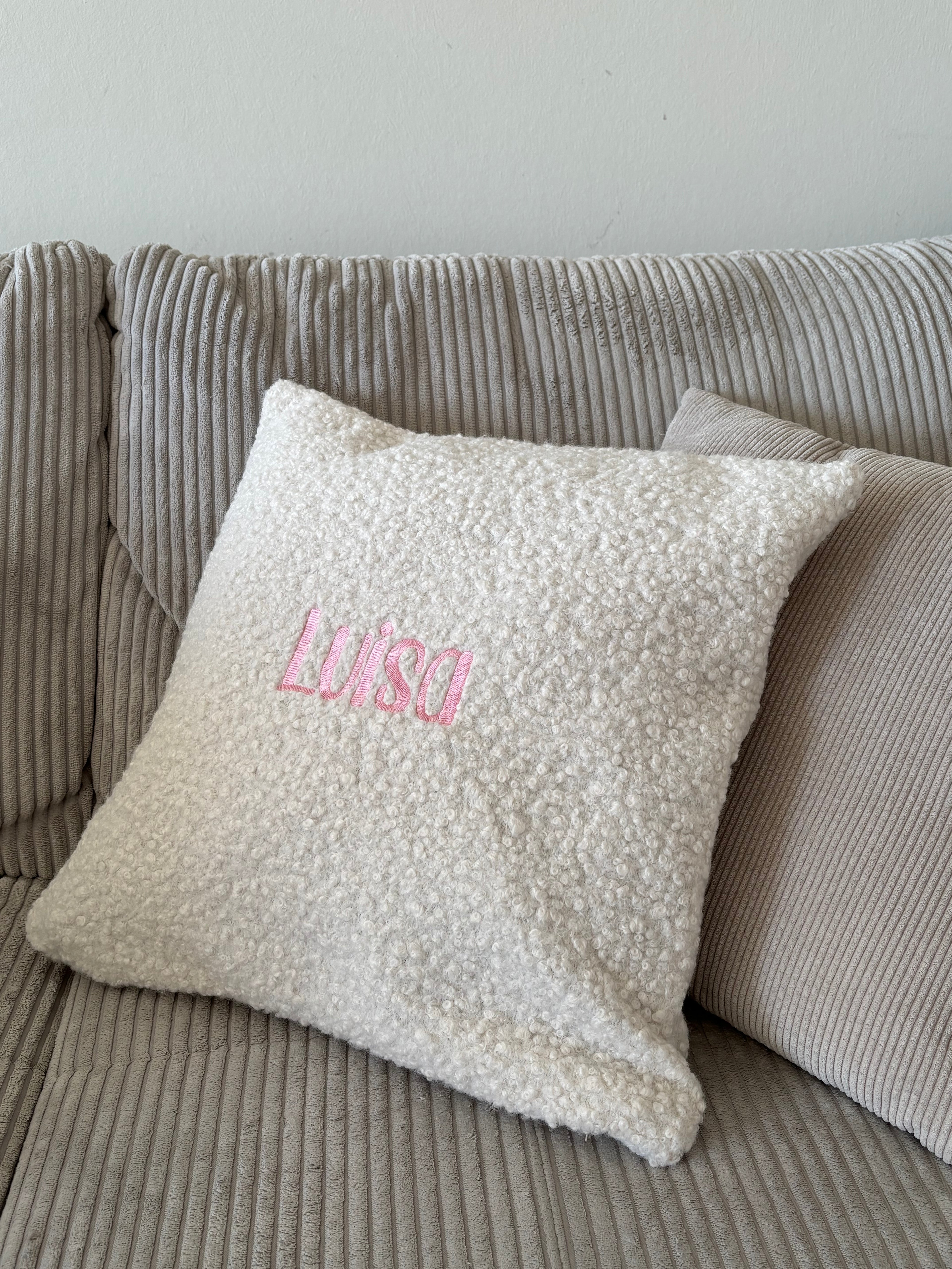 Ein besticktes Kissen aus Boucle Stoff mit einem rosa Wunschtext "Luisa".