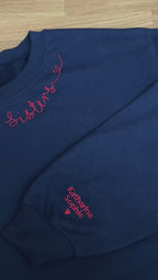 Ein Sweater in der Farbe Navy Blue mit einer roten Stickerei am Kragen: Sisters.