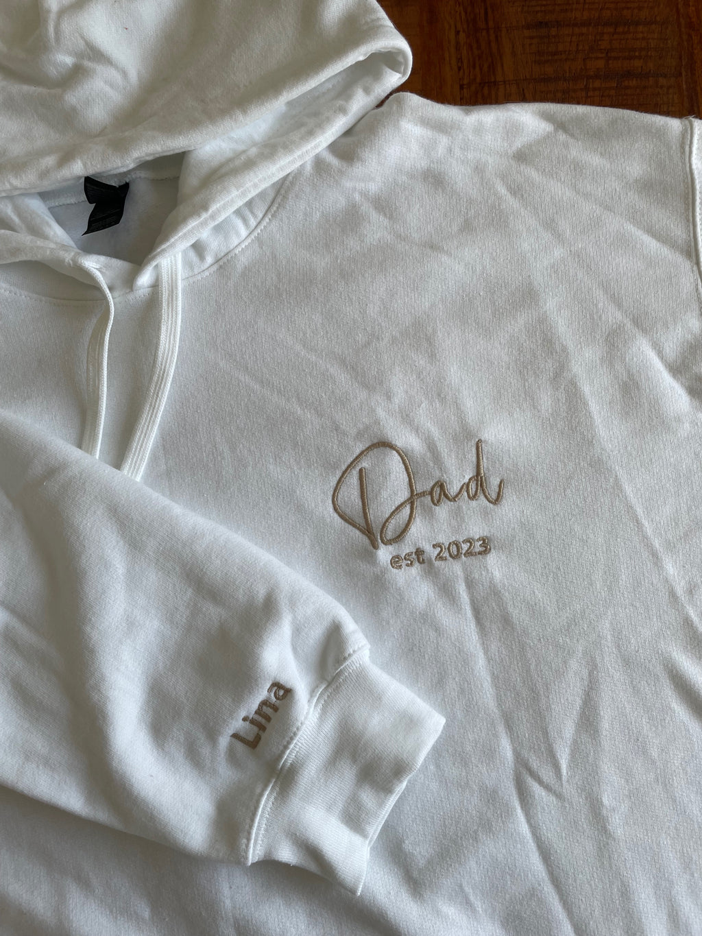 Ein weißer Hoodie mit personalisiertem Text auf der linken Brust. Es ist "Dad est 2023" in beige bestickt und auf dem Ärmel "Lina".
