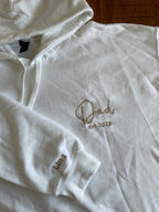 Ein weißer Hoodie mit personalisiertem Text auf der linken Brust. Es ist "Dad est 2023" in beige bestickt und auf dem Ärmel "Lina".