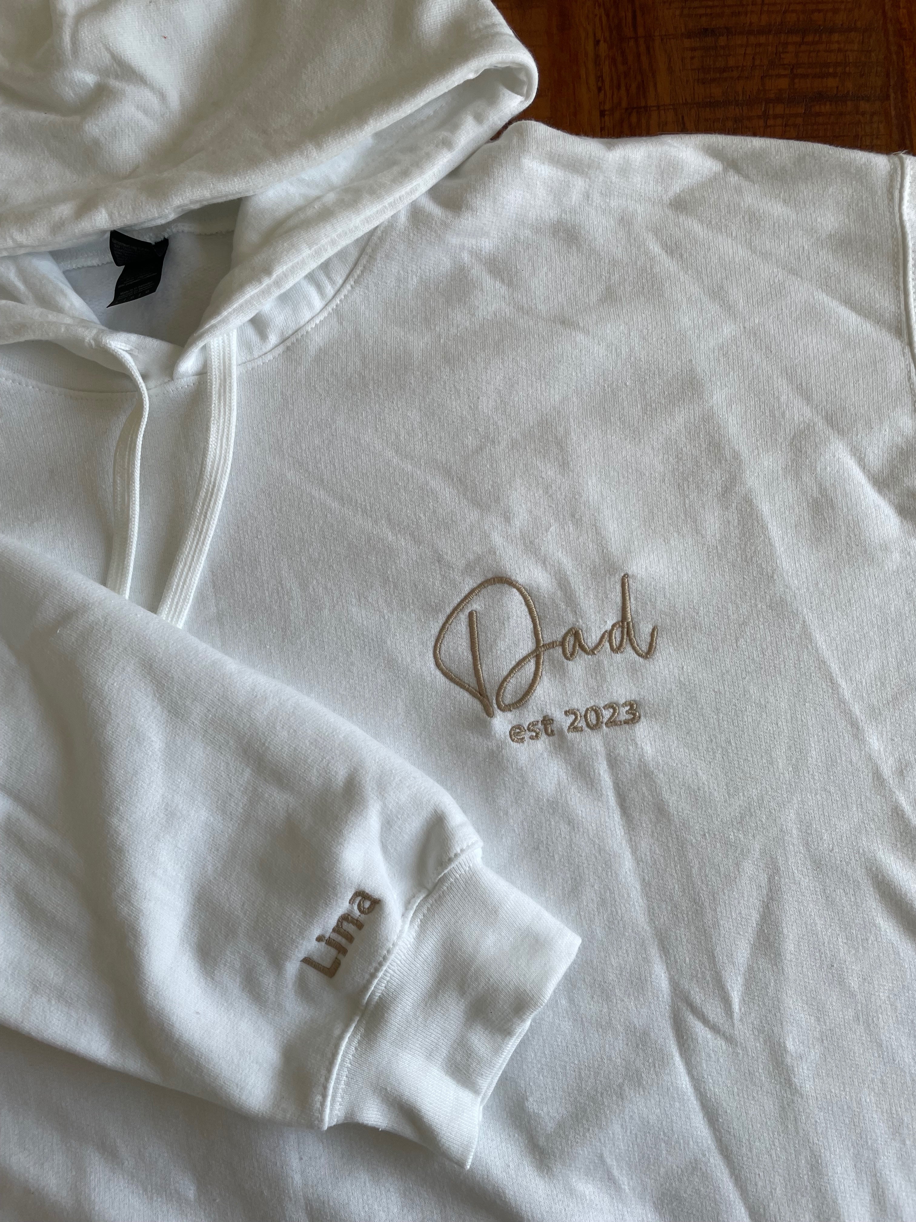 Ein weißer Hoodie mit personalisiertem Text auf der linken Brust. Es ist "Dad est 2023" in beige bestickt und auf dem Ärmel "Lina".