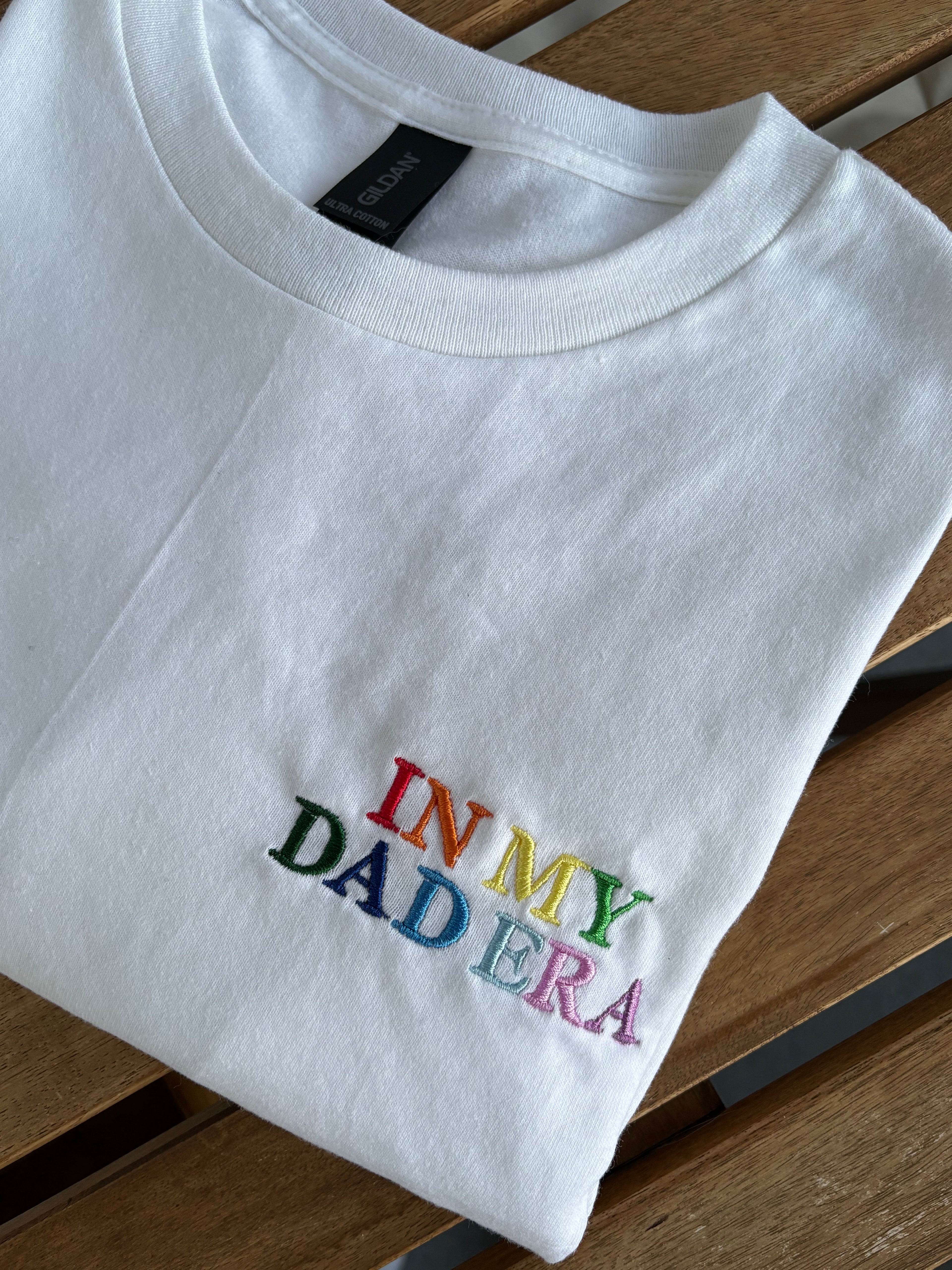 Ein weißes T-Shirt mit einem bunten Stick vorne links auf der Brust "In My Dad Era" in Großbuchstaben. Das Shirt liegt auf einem Holztisch.