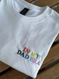 Ein weißes T-Shirt mit einem bunten Stick vorne links auf der Brust "In My Dad Era" in Großbuchstaben. Das Shirt liegt auf einem Holztisch.